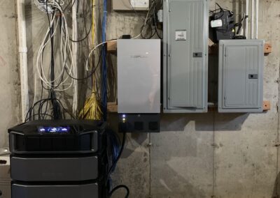 A basement wall with electrical panels, wires, and a modern battery storage system with three stacked units.