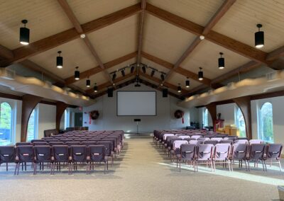A spacious meeting hall with rows of empty chairs facing a large screen and podium, wooden beams overhead, and natural light coming through side windows.