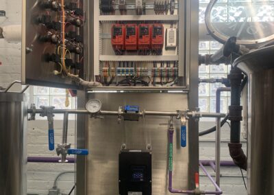 Industrial control panel with wiring and components, flanked by pipes and gauges, in a metal enclosure.